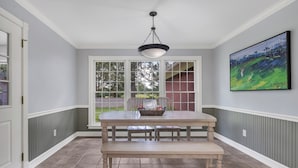 Un coin repas avec une table et un banc, de grandes fenêtres, un luminaire suspendu et un tableau encadré au mur.