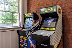 Zwei Vintage-Spielautomaten, einer mit der Aufschrift 'Golden Tee 3D Golf' und der andere 'Arcade 1UP', vor einer Ziegelwand mit einem Fenster im Hintergrund.