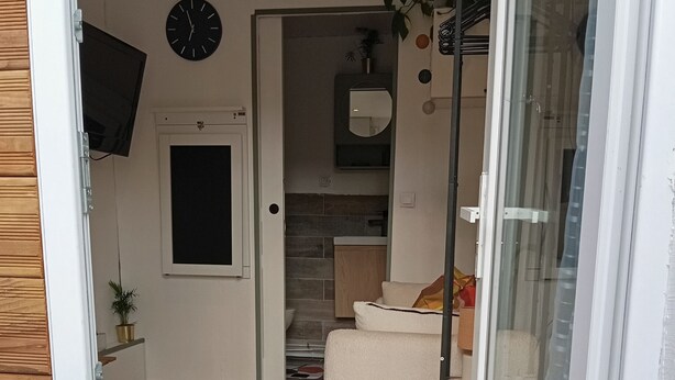 A room with a wooden floor, a white sofa, a glass door, and a kitchen area with a clock on the wall. - Cozy room 15 minutes from Paris-Orly with a bathroom and private terrace (Choisy-le-Roi)