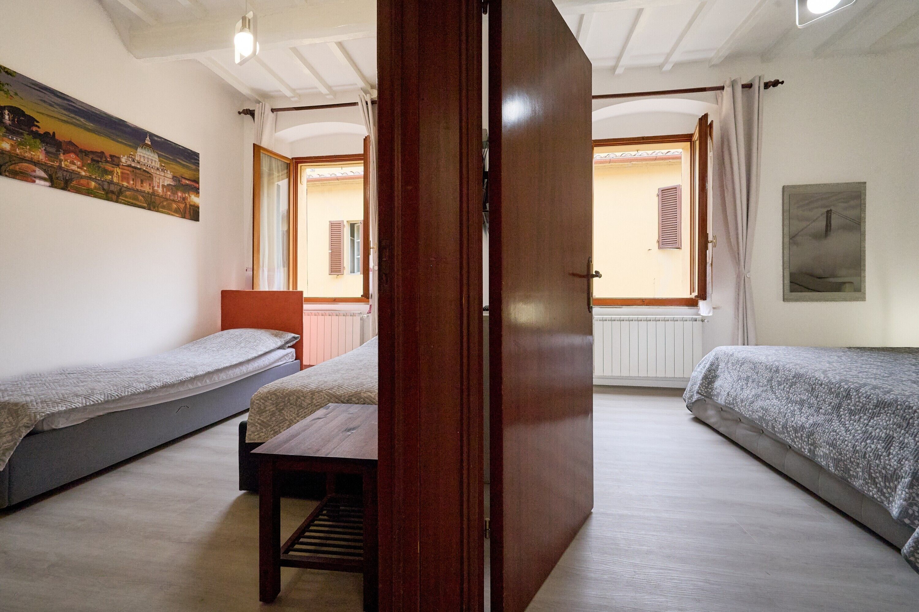 Una habitación con dos camas individuales, una puerta de madera, una mesita y vistas a un edificio desde la ventana.
