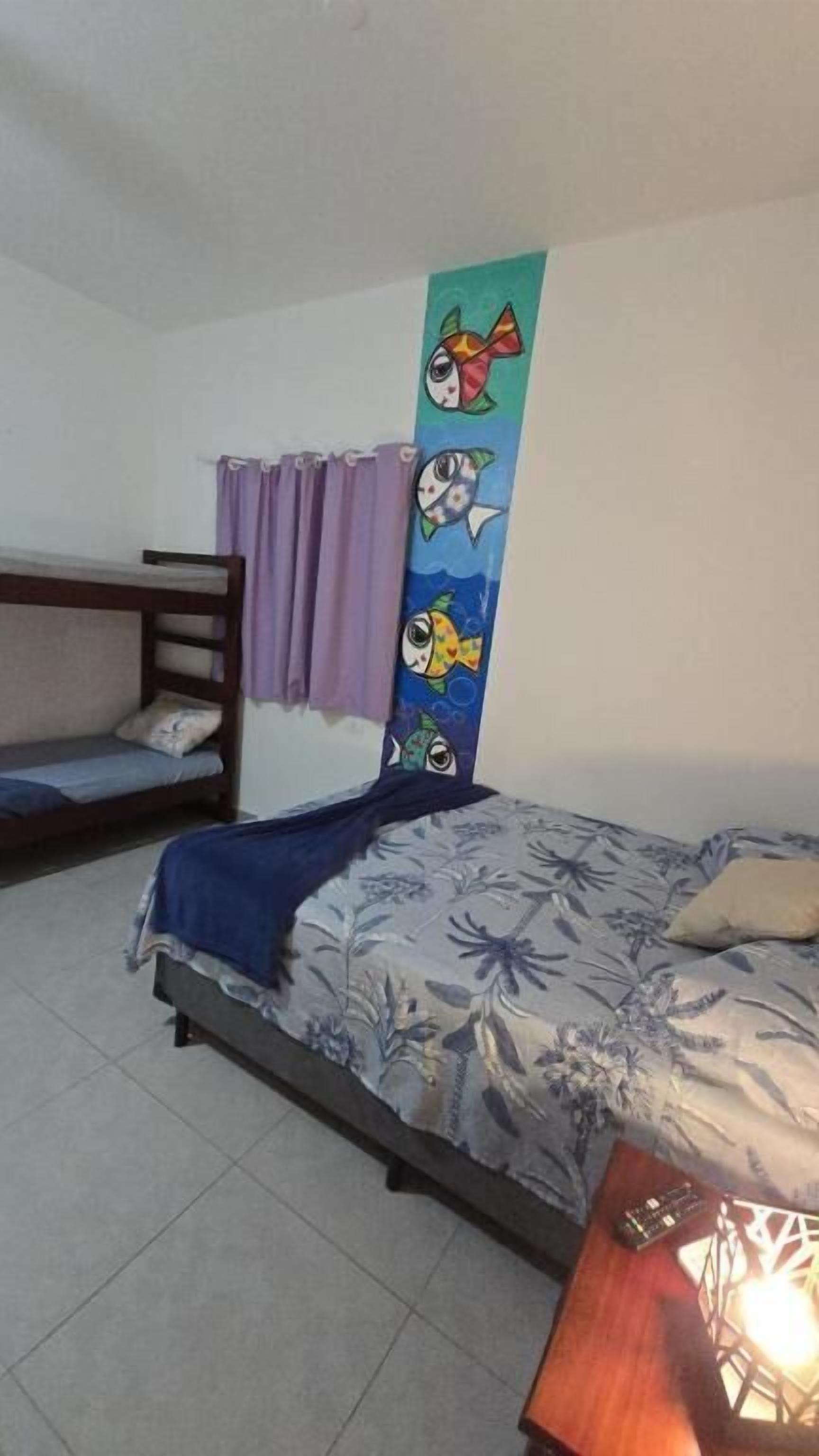 Um quarto com cama, beliche, parede com mural de peixes coloridos, cortina e mesa de cabeceira com luminária.