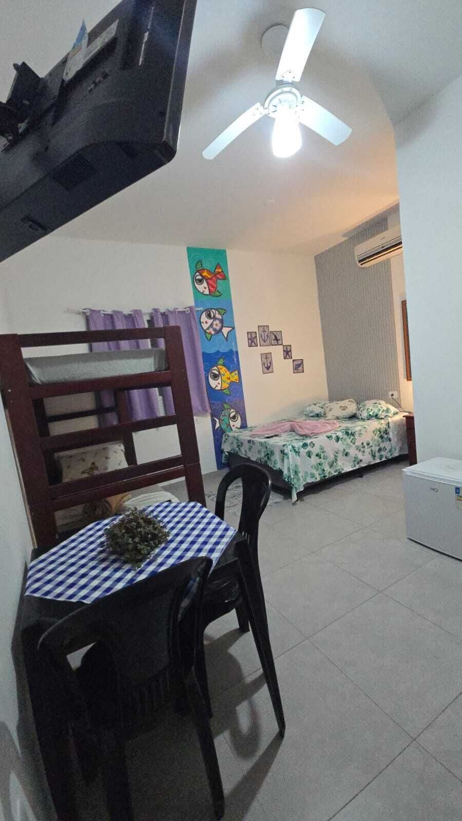 A room with a bed, a table with a checkered tablecloth, a ceiling fan, and a wall with drawings.