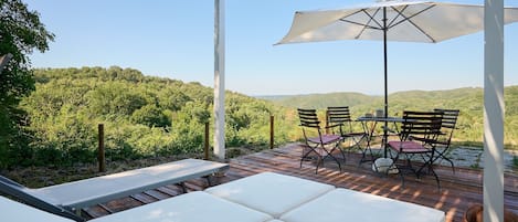 A covered outdoor seating area with a table and chairs, a large umbrella, and a view of a forested hillside.