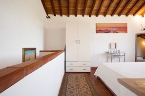 Une chambre avec un plafond en bois, un lit recouvert d’une courtepointe blanche, une table de chevet, une petite table avec une horloge, une armoire et un tableau représentant un coucher de soleil.
