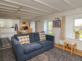 A living room with a blue sofa, a small wooden table, a patterned rug, a kitchen in the background, and a window with curtains.