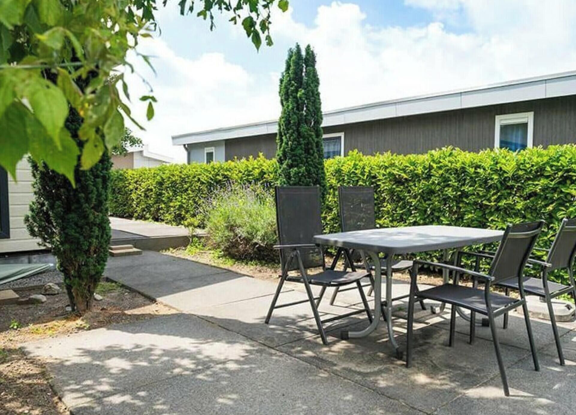 Um pátio externo com mesa e cadeiras, um sebe e um edifício ao fundo.