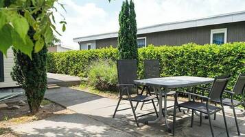 Um pátio externo com mesa e cadeiras, um sebe e um edifício ao fundo.