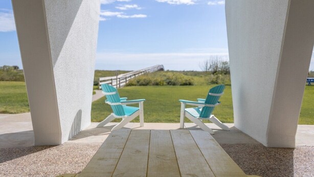 A covered porch with two turquoise Adirondack chairs, a wooden table, and a view of a grassy area with a wooden walkway. - 218 Harbor View Studio for Two Pool & Beach (Galveston)