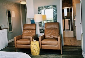 Une chambre d’hôtel avec deux fauteuils en cuir, un petit pouf jaune, un lit, une grande fenêtre et une salle de bain visible à travers une porte ouverte.