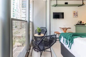Ein kleiner, moderner Raum mit einem Bett, einem Fernseher, einem Tisch mit einer Pflanze und einem Balkon mit Blick auf Gebäude.