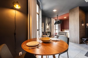 A dining area with a round wooden table, plates, cutlery, and a bowl. There is a wall-mounted lamp, a flat-screen TV, and a sofa in the background. - Jp047 Jacques Pilon Residence 3 Pessoas (São Paulo)
