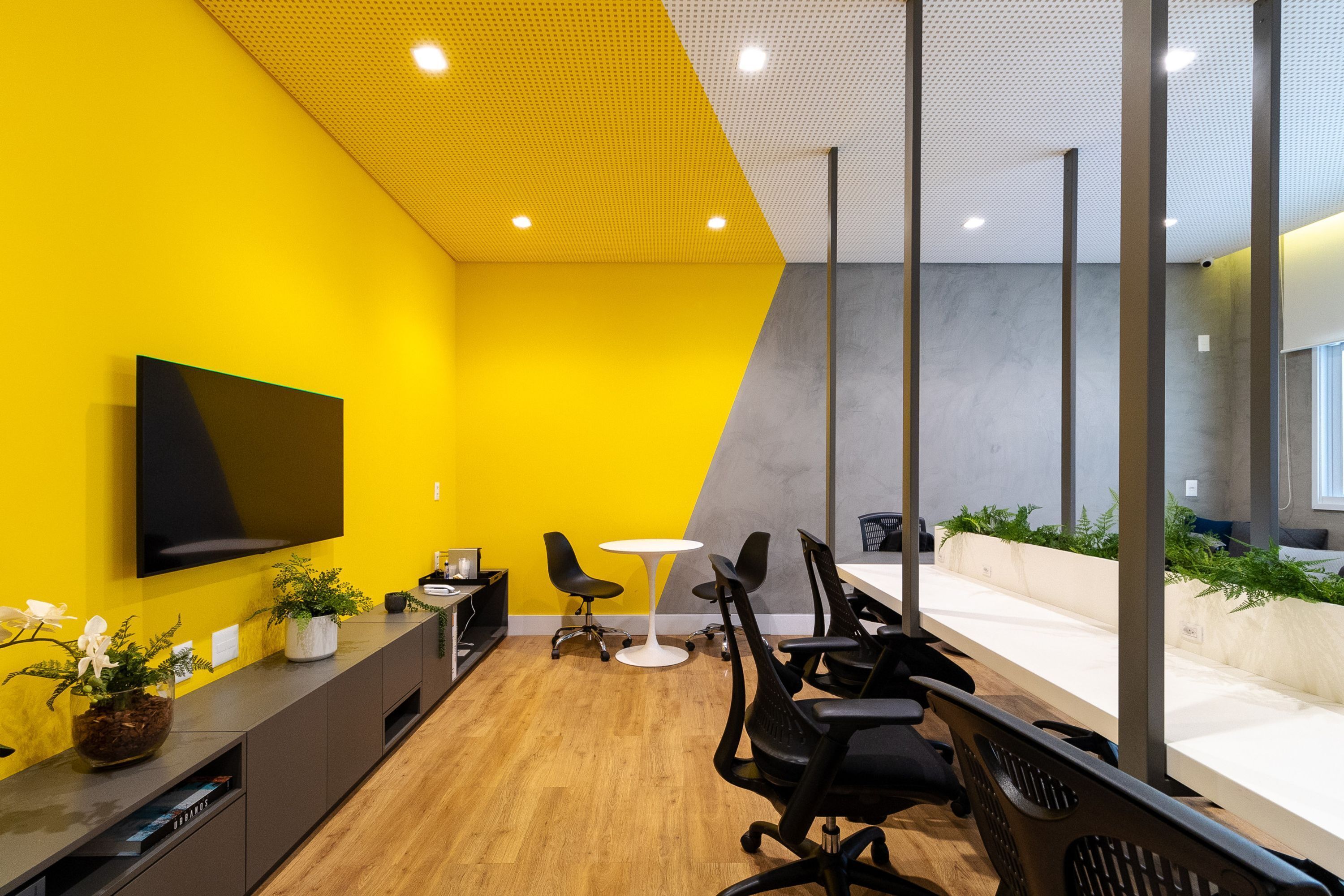 A modern office space with a yellow accent wall, a flat-screen TV, black office chairs, a white table, and potted plants.