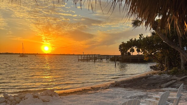 Escena de playa con atardecer, sillas y un muelle.
