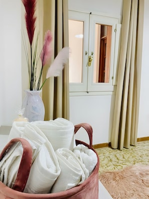 A room with a window, a vase with dried plants, and a basket of towels.