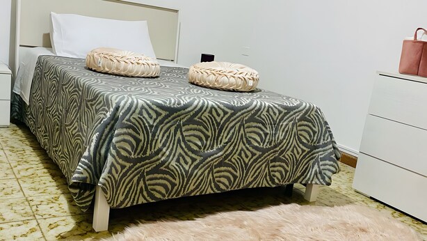 A bed with patterned bedding, a fluffy rug, a bedside table, and a dresser with a bag on top.