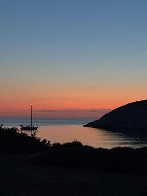 Un coucher de soleil sur une mer calme, avec un voilier et la silhouette d’une colline.