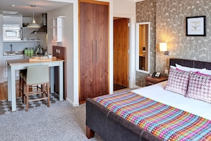 A hotel room with a bed, bedside table, lamp, and a desk with a chair. There is a wardrobe and a door leading to another room.