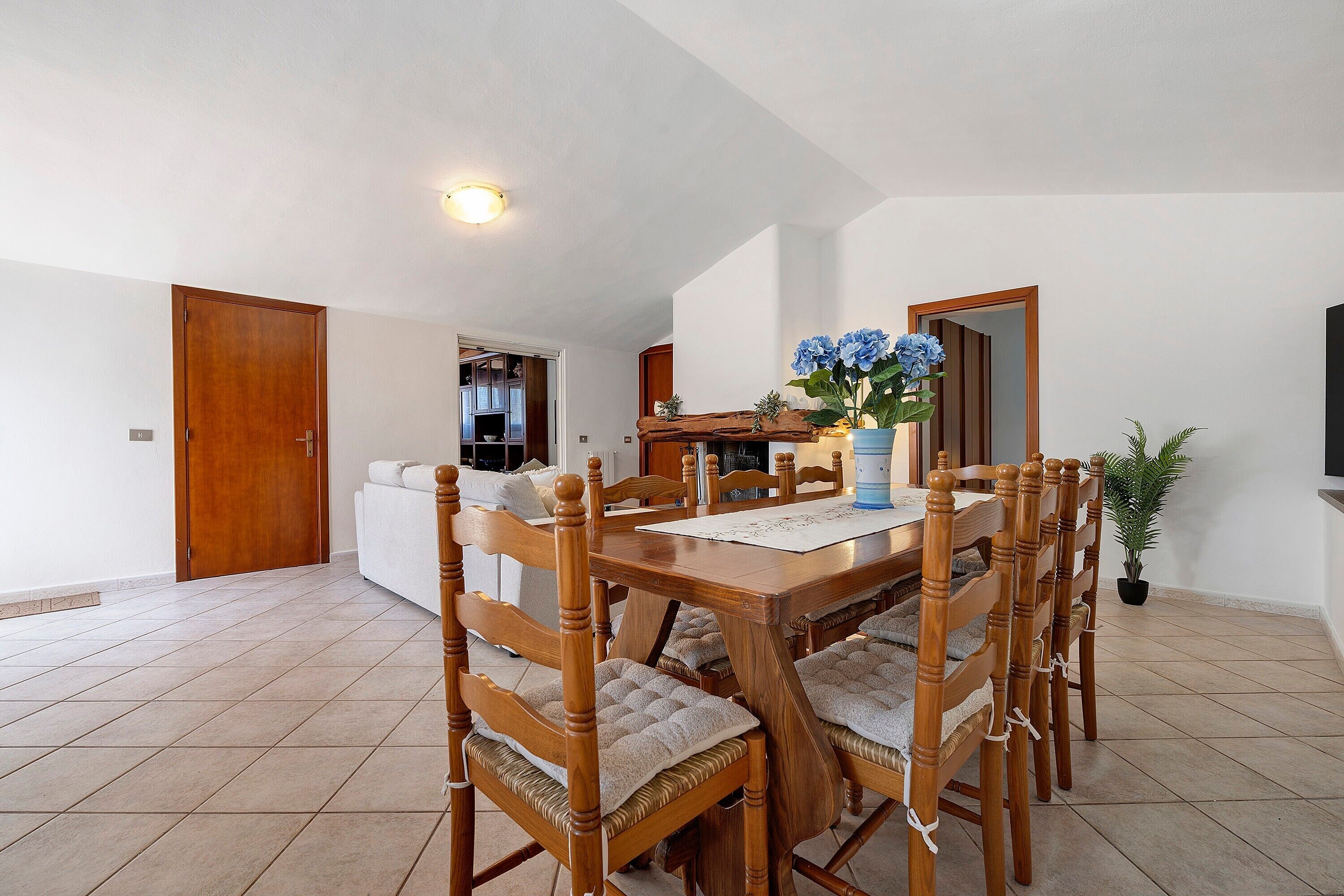 A dining area with a wooden table and chairs, a vase with flowers, and a view into the kitchen.