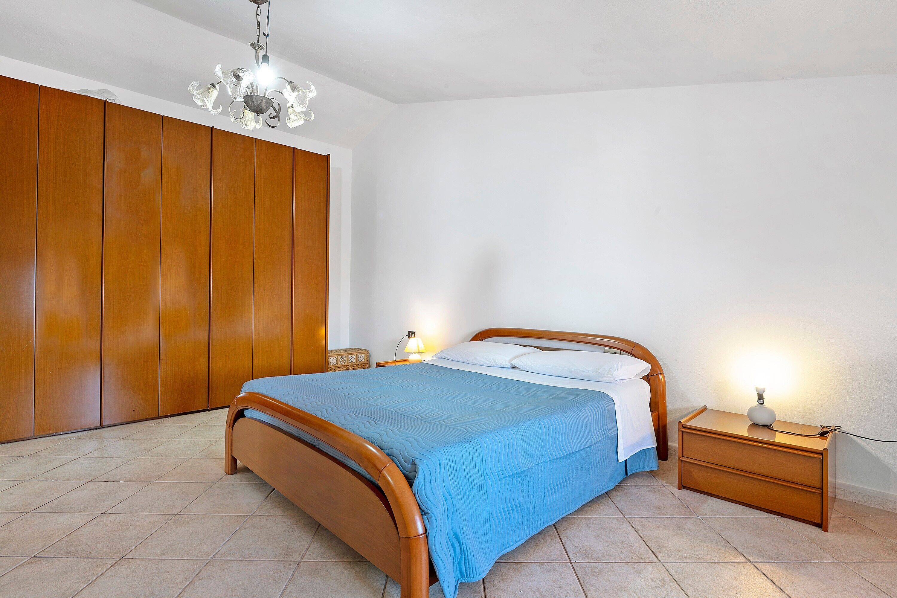 Ein Schlafzimmer mit einem hölzernen Bett, einem Nachttisch mit Lampe, einem Kleiderschrank und einer Pendelleuchte.