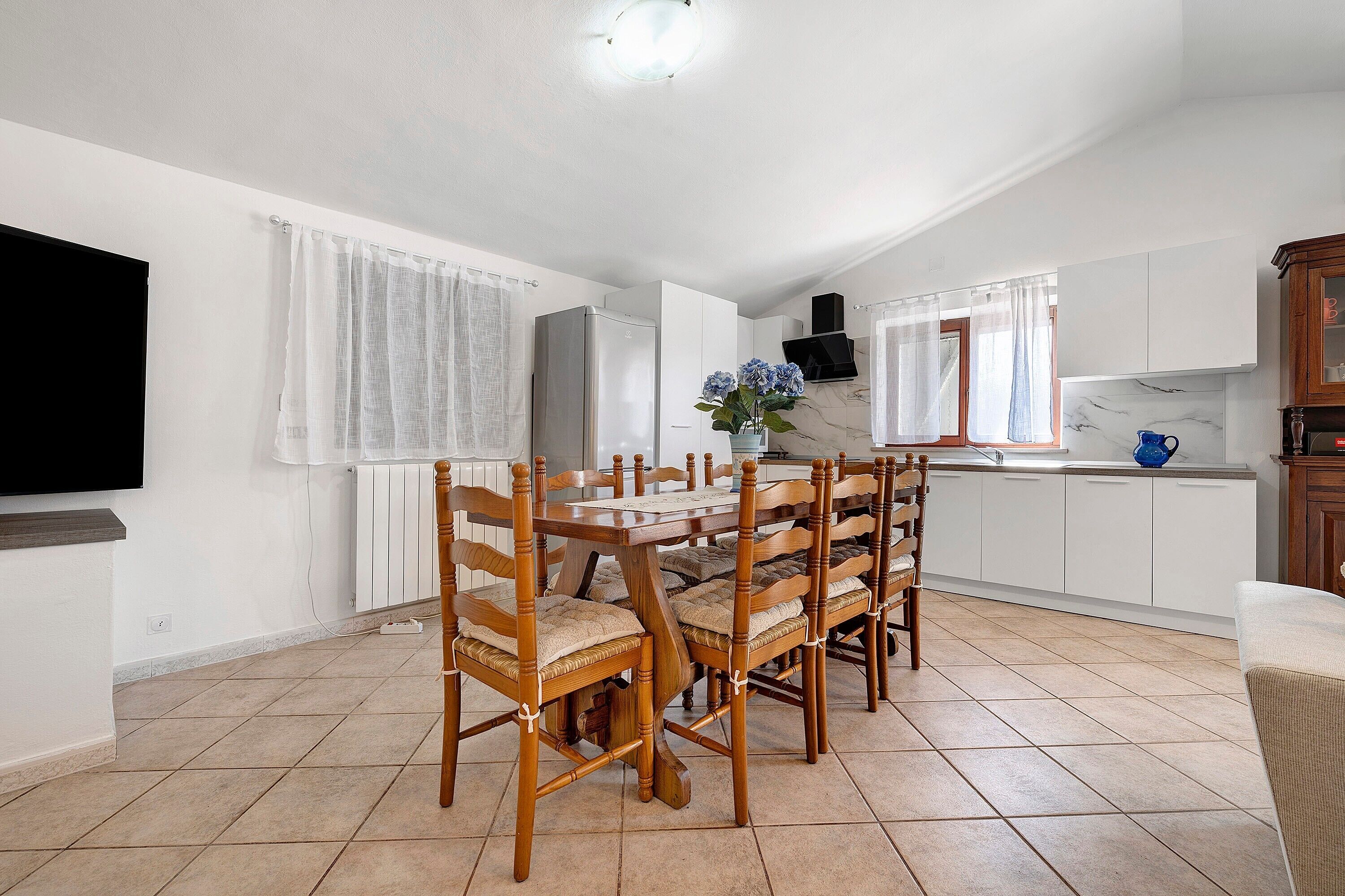 Eine moderne Küche mit weißen Schränken, einem Esstisch mit Holzstühlen und einem an der Wand befestigten Fernseher.