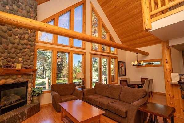 A cozy living room with a stone fireplace, wooden beams, large windows, and a dining area.