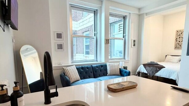 A modern kitchen with a sink, countertop, and a dishwasher. There is a blue sofa with a window view and a bed in the adjacent room.