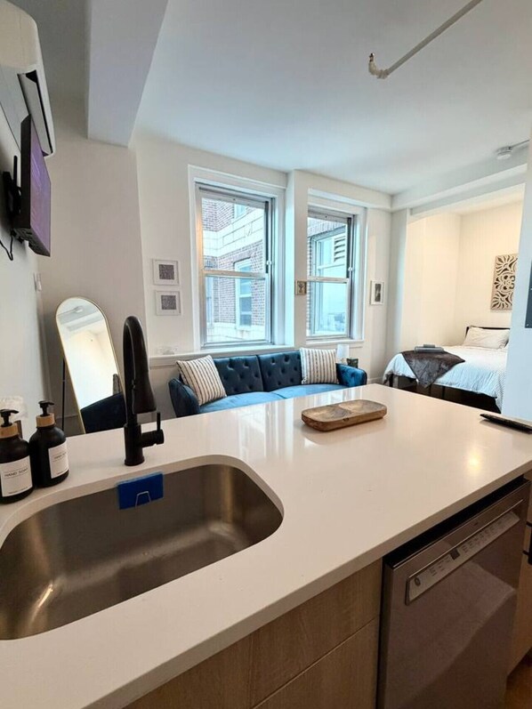 A modern kitchen with a sink, countertop, and a dishwasher. There is a blue sofa with a window view and a bed in the adjacent room.