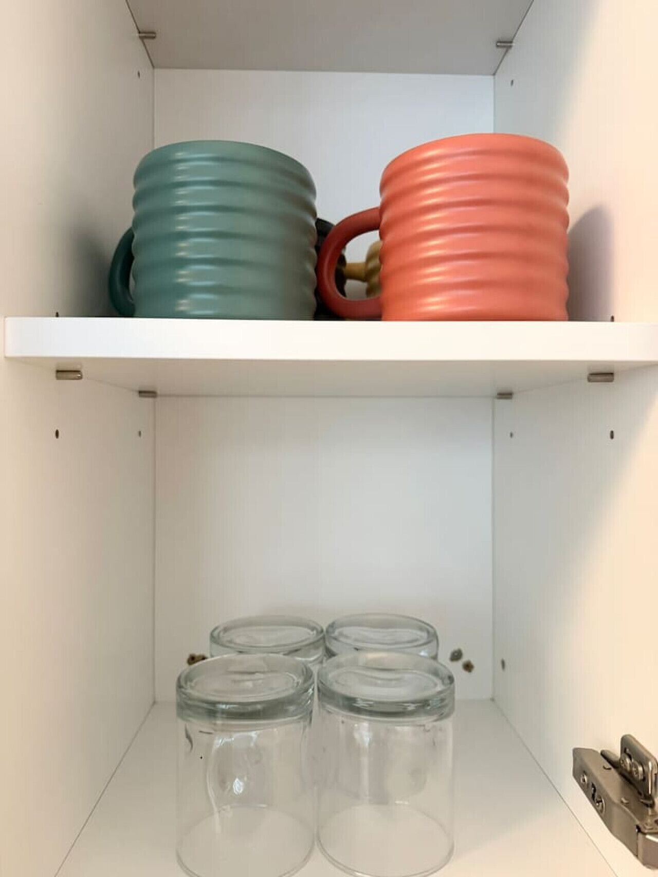 A white shelf with two colorful mugs and three clear glass jars.