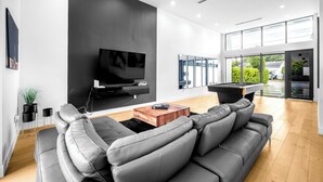 A modern living room with a large gray sofa, a flat-screen TV mounted on a black wall, a wooden coffee table, and a pool table. - Noir Luxury Home, Design District w/ Pool, Billiard (Miami)