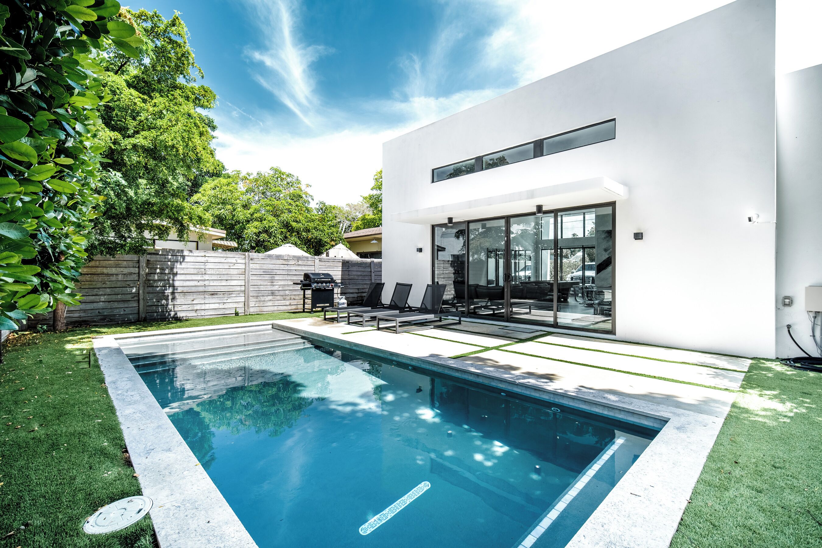 Une maison moderne dotée d’une piscine, d’un patio avec des chaises longues et d’une clôture en bois.