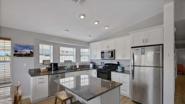 A modern kitchen with white cabinets, a stainless steel refrigerator, a microwave, and a granite countertop island with stools. - Steps to Beach Walk to Pier, Restaurants & Nightlife Near Downtown & Amphitheatre (St. Augustine)