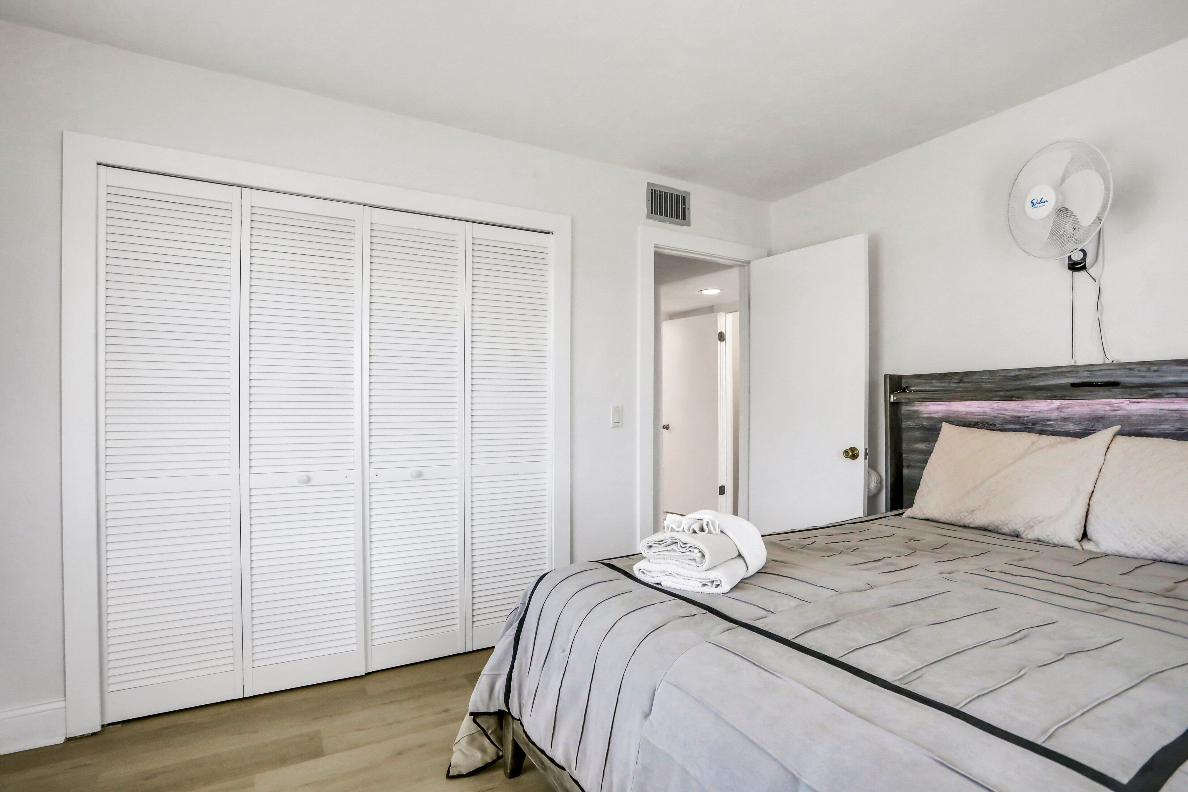 A bedroom with a bed, a white closet, a fan, and a door leading to another room.