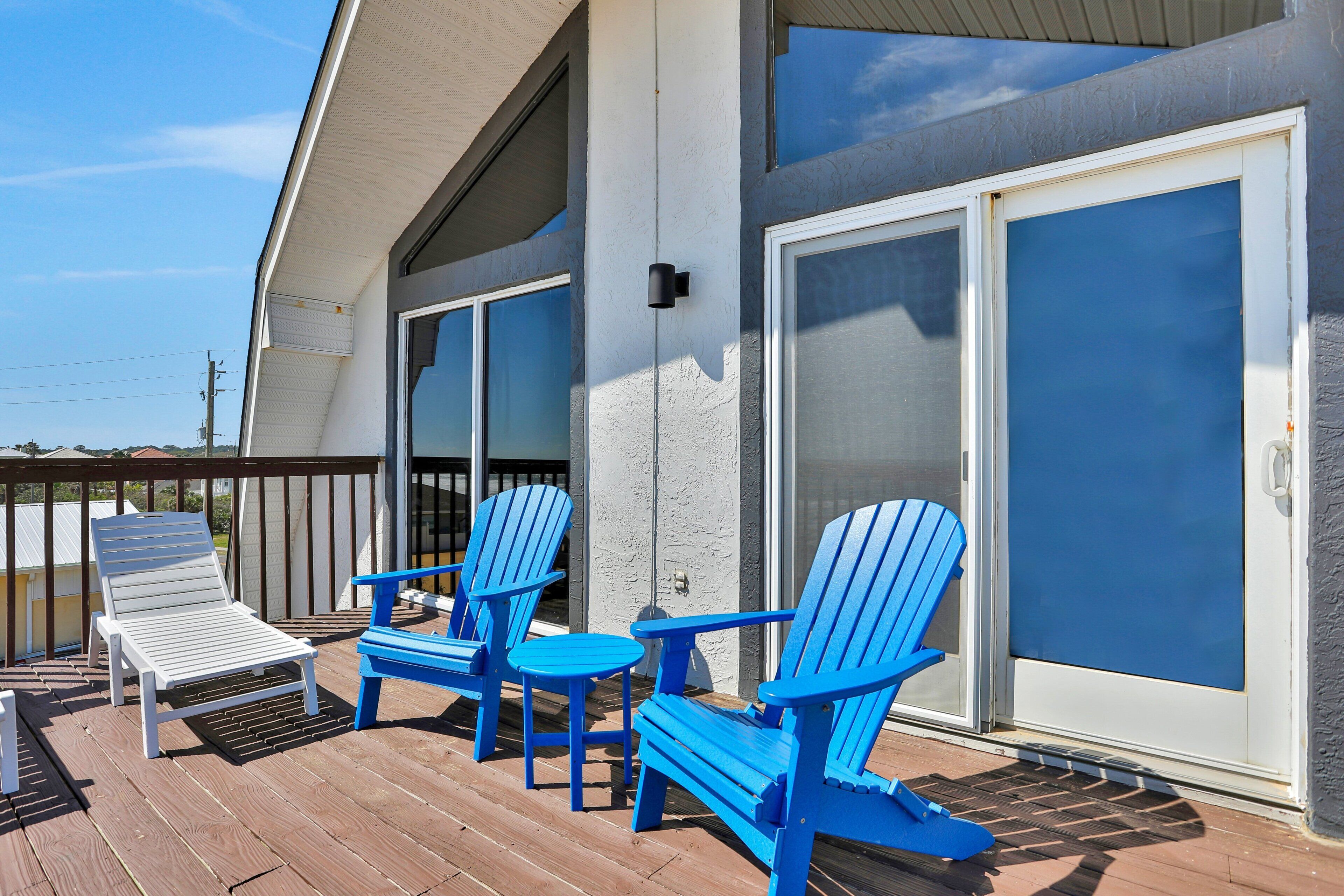 A deck with blue Adirondack chairs and a white lounge chair, a sliding glass door, and a white exterior wall.