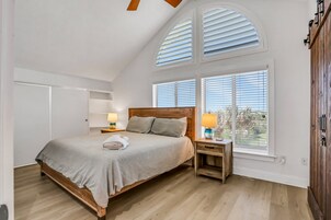 A bedroom with a wooden bed, two bedside tables with lamps, a large window with blinds, and a view of the outdoors.
