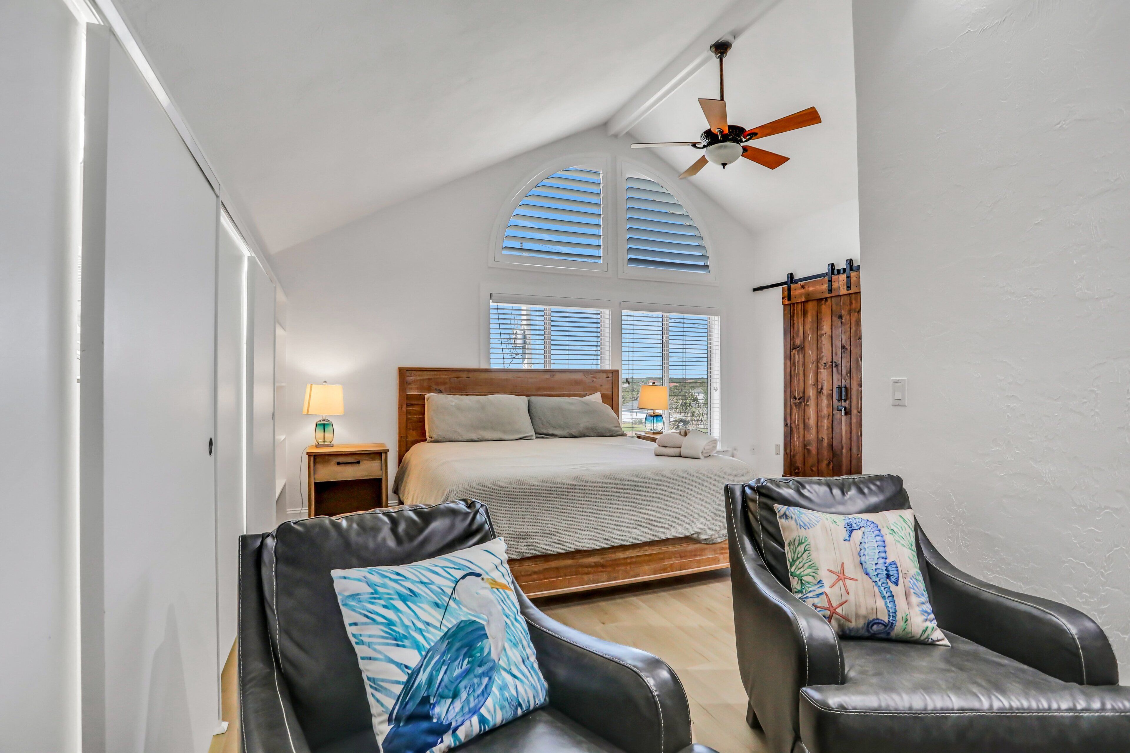 A bedroom with a bed, two chairs, a ceiling fan, and a wooden door.