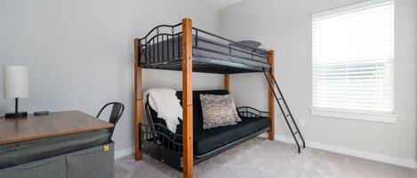 Een stapelbed met een bureau en stoel, een raam met zonwering, en een vloerbedekking.
