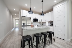 Una cocina moderna con armarios blancos, un refrigerador de acero inoxidable, un microondas y un lavamanos. Hay tres taburetes negros y lámparas colgantes sobre la encimera.