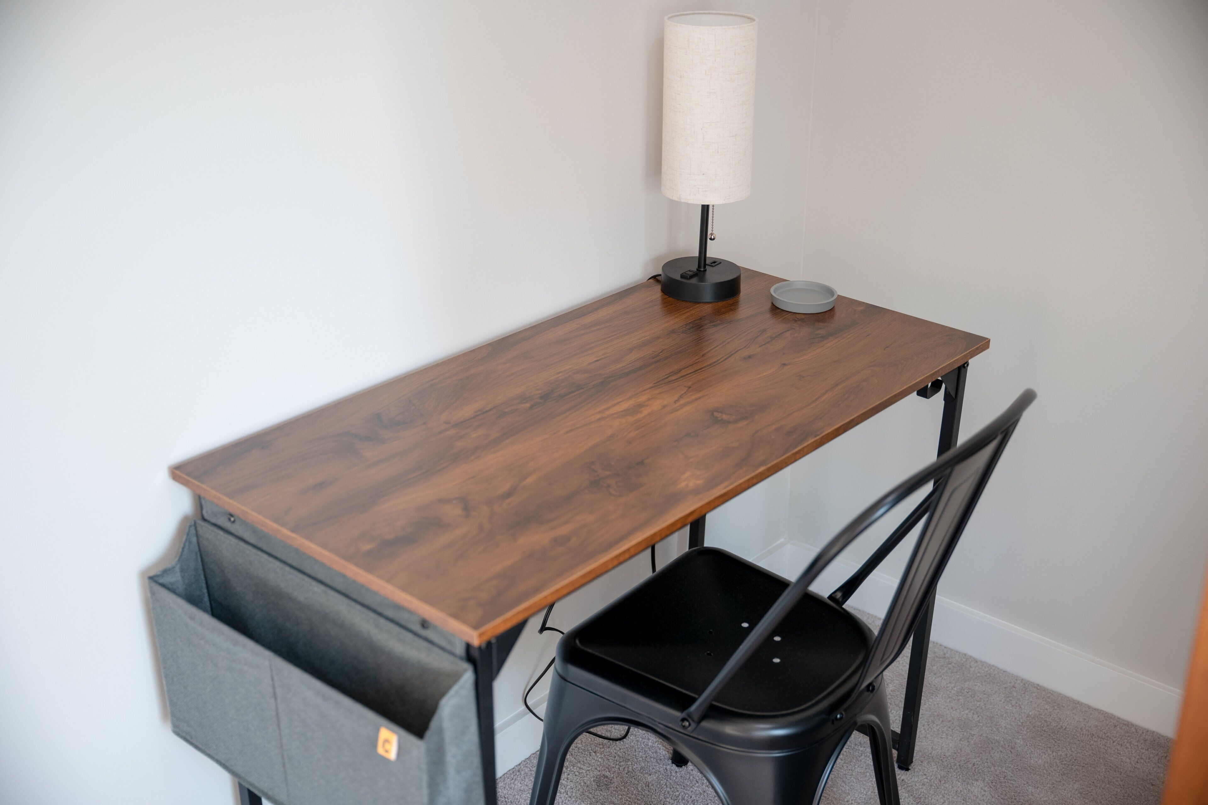Un escritorio de madera con una lámpara, una silla negra y un contenedor gris.