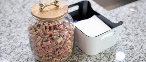 A glass jar with a wooden lid and a metal handle, filled with small, pink and beige items, next to a white container with a black handle on a granite countertop.