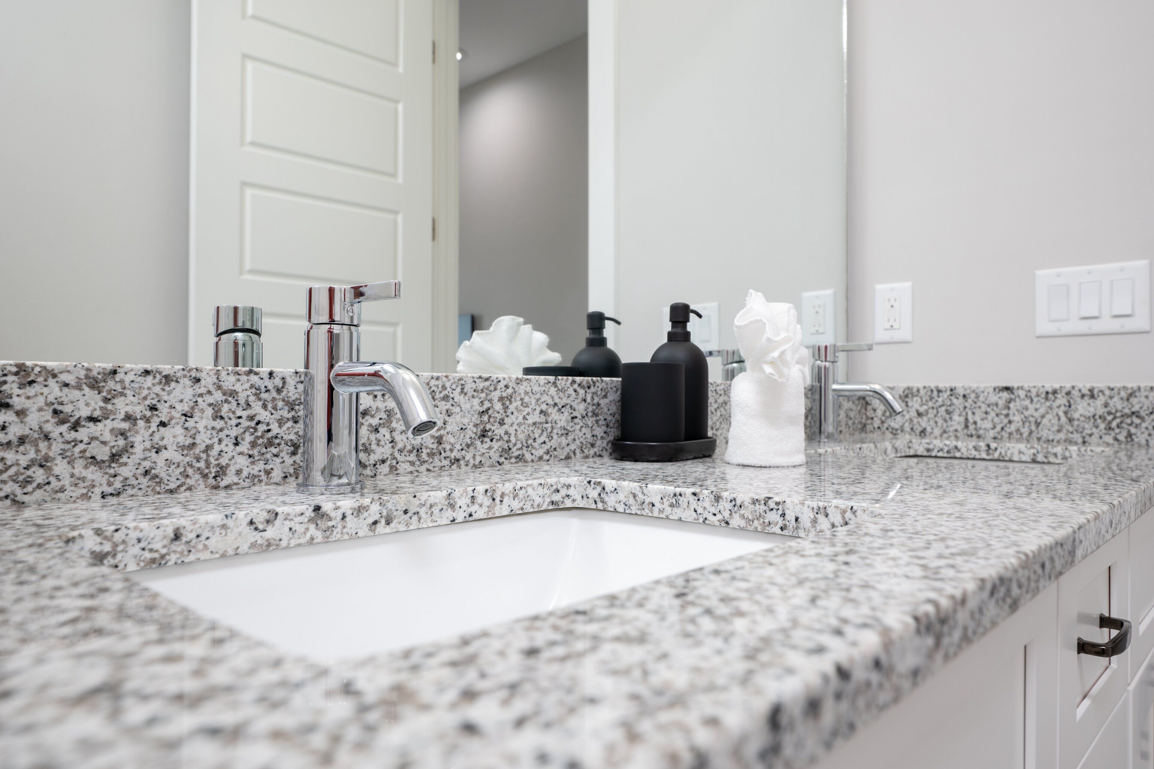 A bathroom with a granite countertop, a white sink, a mirror, a door, and a towel dispenser.