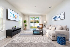 A modern living room with a flat-screen TV, a sofa, a coffee table, and a large window with a view. - Pet-friendly 4BR Beach Home Walk to Beach & Jacksonville Beach Pier Fenced Yard (Jacksonville Beach)