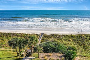 A beach view with a wooden walkway leading to the shore, surrounded by palm trees and vegetation. - Oceanfront Luxury Condo Renovated Private Beach Access Pools Tennis Court (St. Augustine)