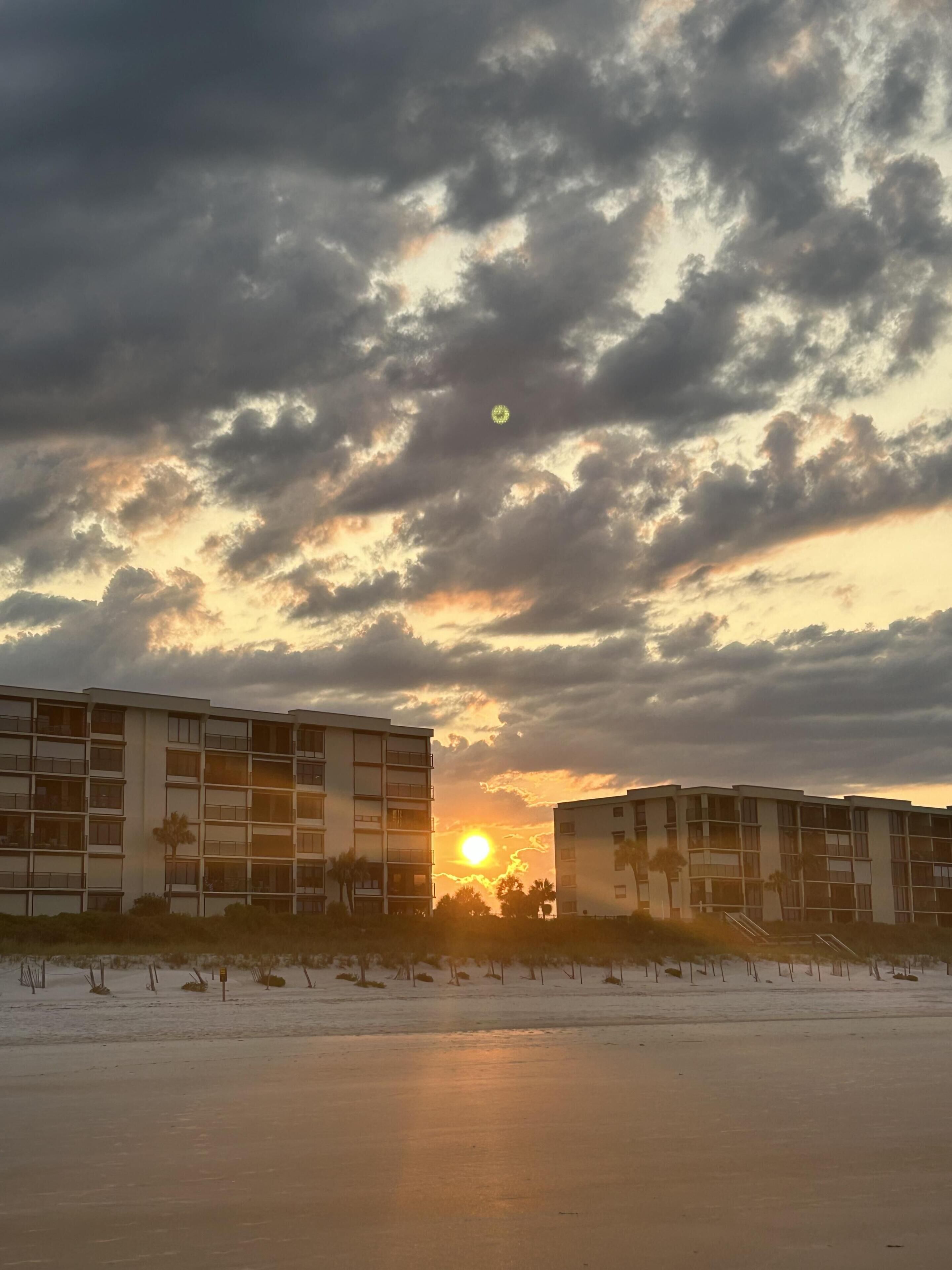 A building with multiple floors and balconies, situated near a beach with a sunset in the background.