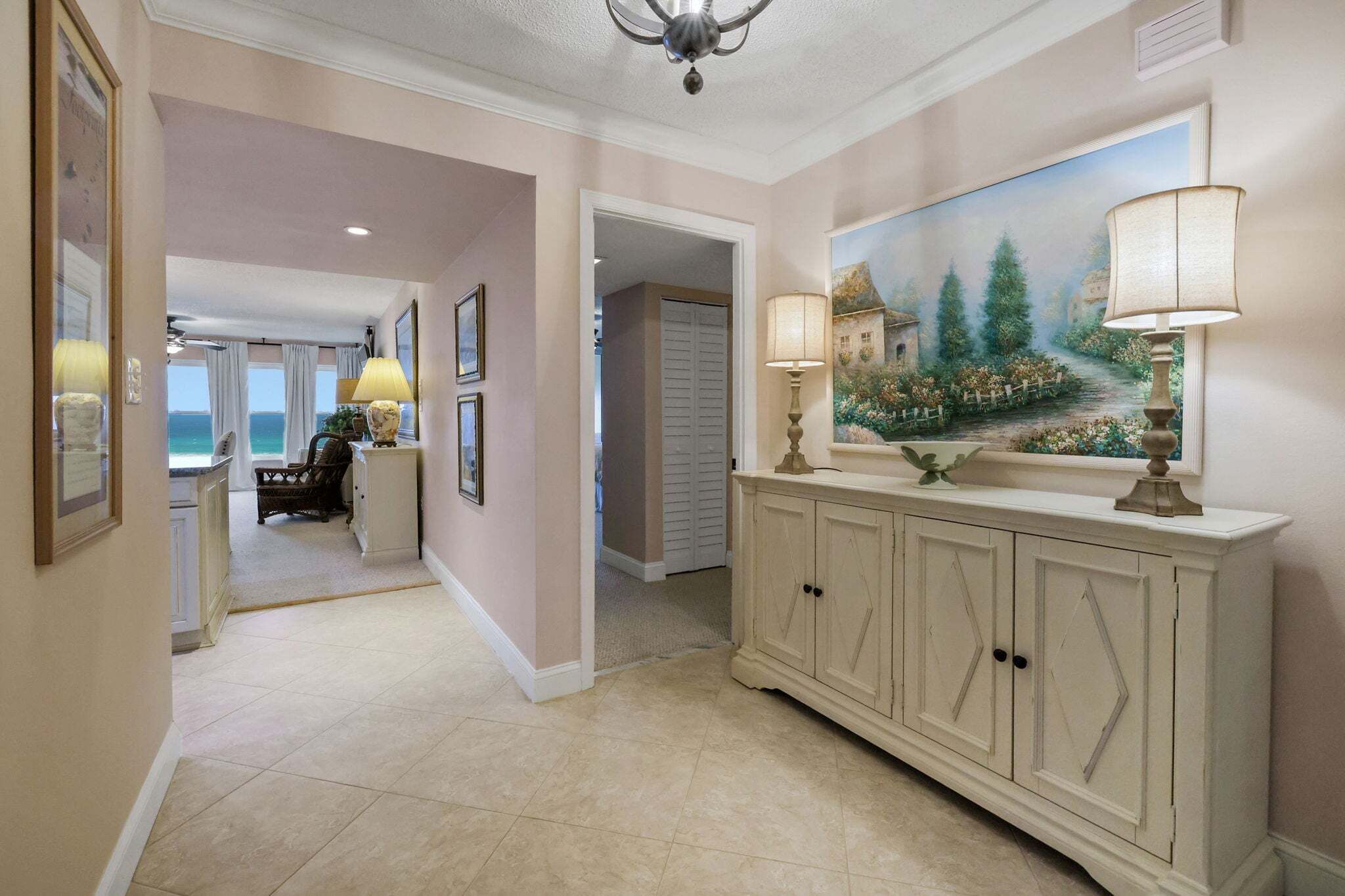 A hallway with a cream-colored floor, a white console table with lamps, a painting of a landscape, and a view to a room with a view.