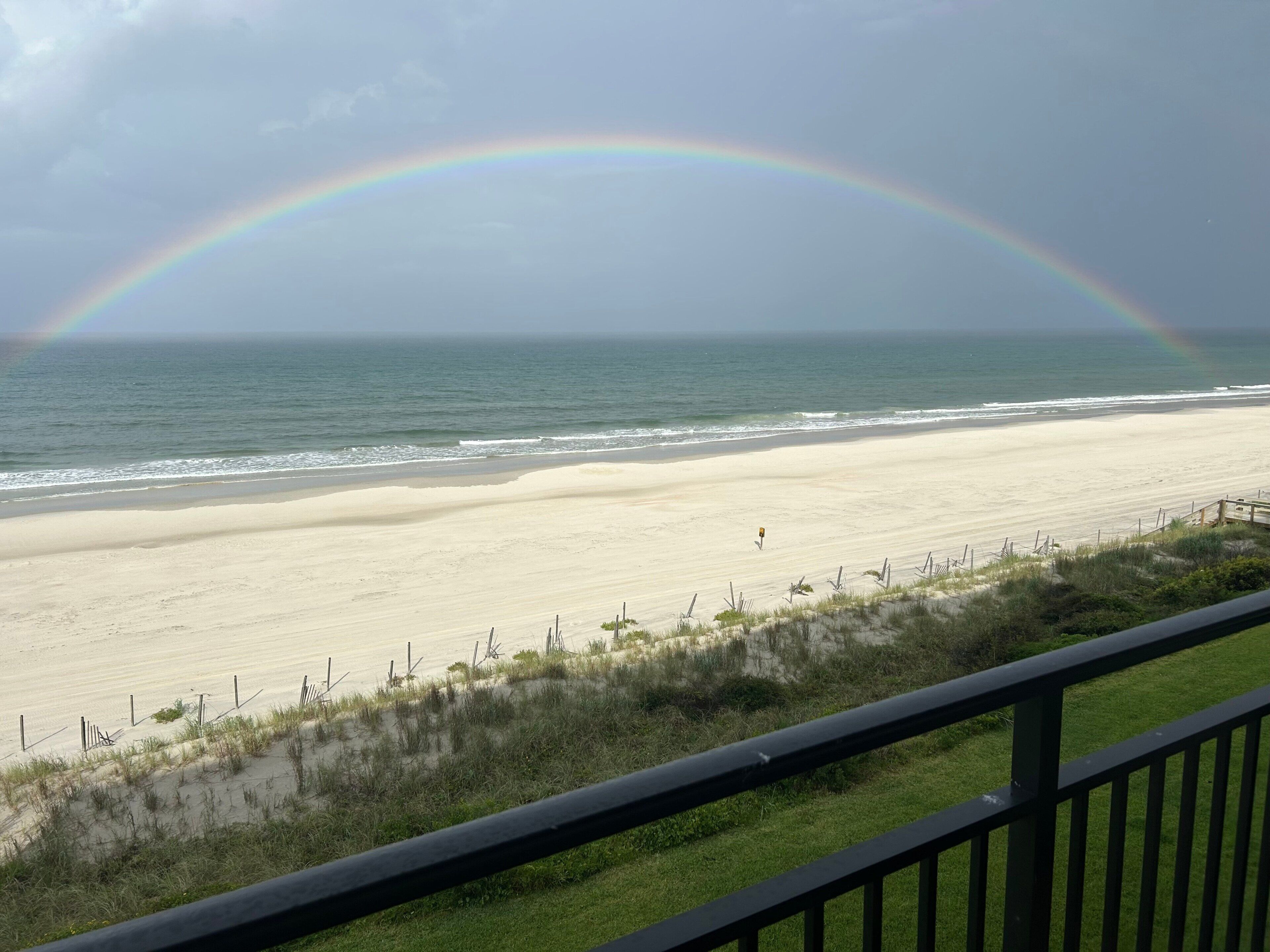 A beach with a rainbow, ocean, and a grassy area with a fence.