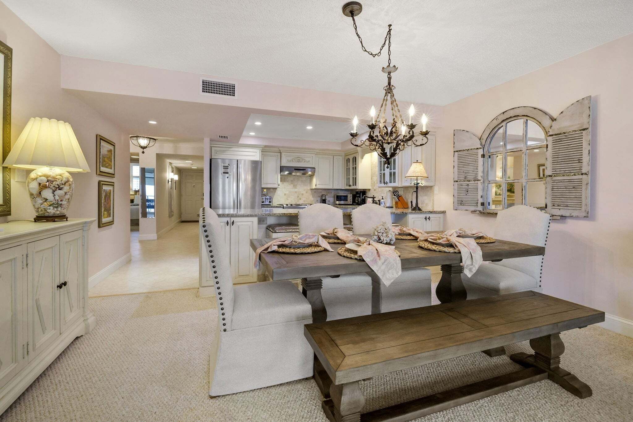 A dining area with a wooden table, cushioned chairs, a chandelier, and a kitchen in the background.