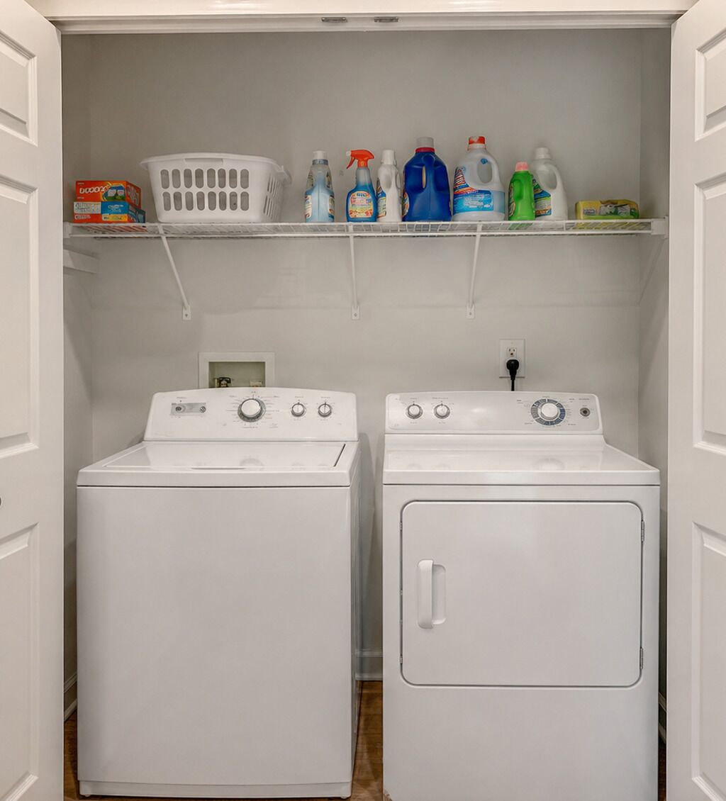 A laundry room with two white front-loading washers and dryers, a basket, and various cleaning supplies on shelves.
