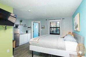 A bedroom with a bed, a bedside table, a window with curtains, a kitchen area with a fridge, and a TV mounted on the wall. - 1915 Beach Club - Lagoon Suite 5 (Daytona Beach)