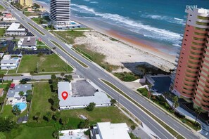 An aerial view of a coastal area with a beach, roads, and buildings including a hotel with a pool and a parking lot.