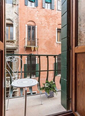 Ein Balkon mit einem Metallstuhl, einem Topfpflanzen und Blick auf ein Gebäude mit grünen Rollläden und Balkonen.